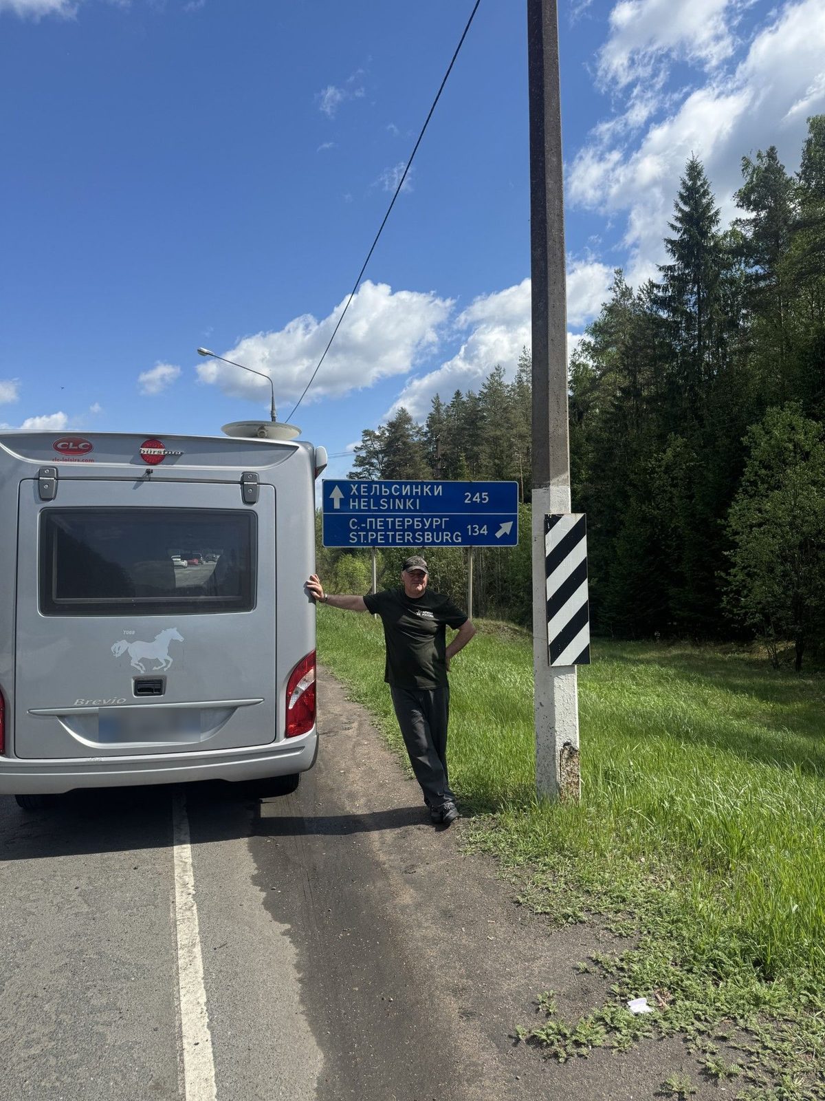 Mirko pose à côté de son camping-car au bord d'une route russe, sous un panneau indiquant Helsinki et Saint-Pétersbourg