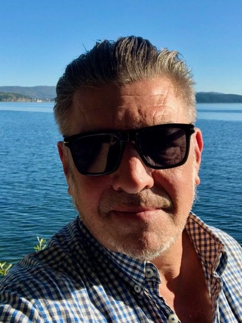 Fabrice, en lunettes de soleil et chemise à carreaux, devant un grand lac bleu en Russie
